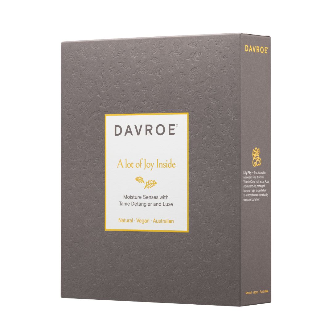 A rectangular product box for DAVROE, shown at a slight angle to display both the front label and the side panel. The box is a textured dark gray with a central white label bordered in gold, featuring the text 'DAVROE', 'A lot of Joy Inside', and 'Moisture Senses with Tama Detangler and Luxe'.