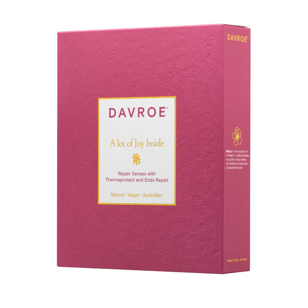 A rectangular pink product box for Davroe 'A lot of Joy Inside' shown at a slight angle on a plain white background. The front has a cream label with gold and dark pink text, and the side panel shows additional text and a small gold icon.