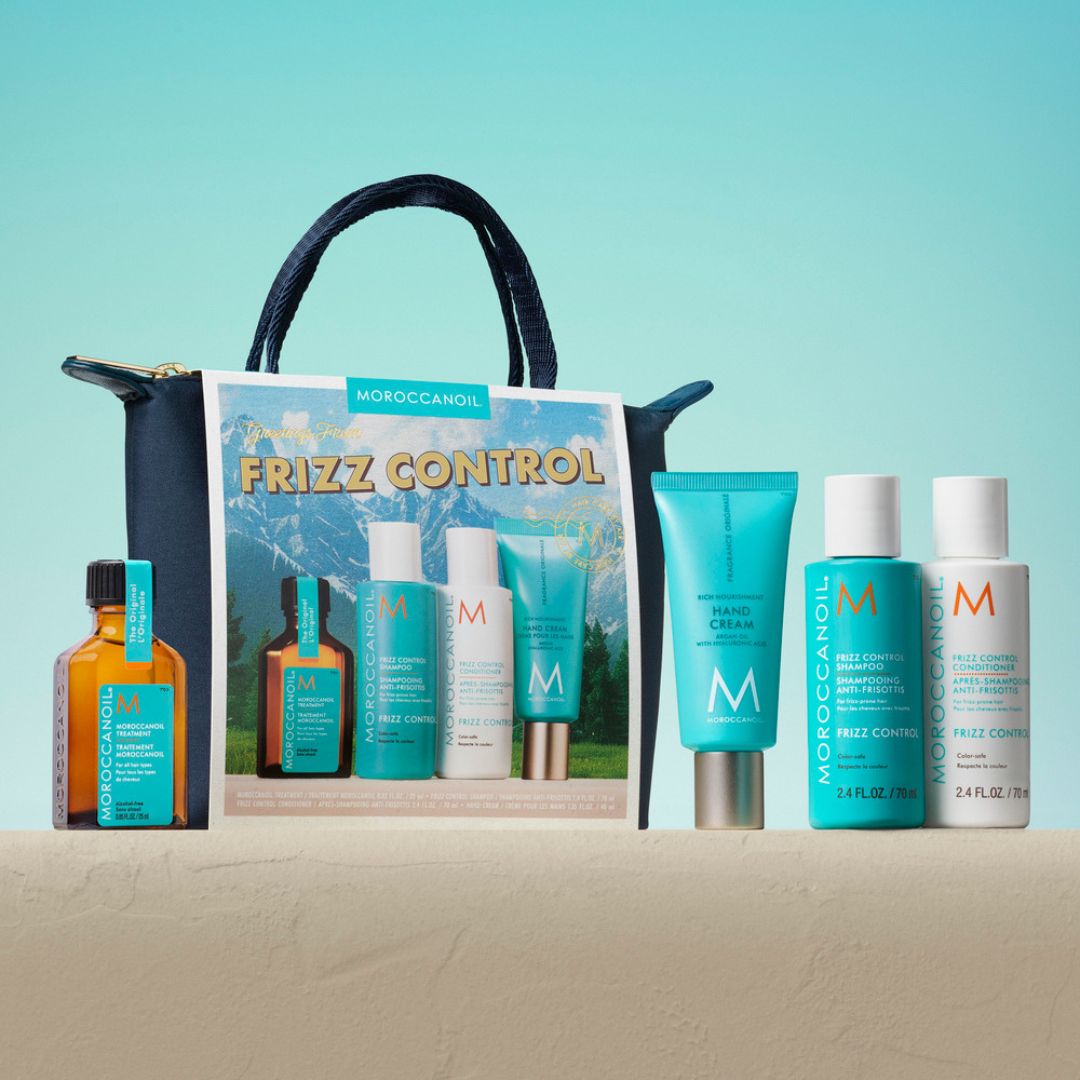 Moroccanoil Frizz Control gift set arranged in front of a branded tote bag on a beige surface against a turquoise gradient background. The set includes an amber bottle, a large turquoise hand cream tube, a turquoise leave-in conditioner bottle, a white treatment bottle, and a white shampoo bottle, with the tote bag behind them.