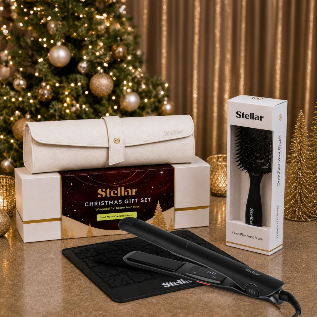 Stellar Christmas gift set featuring a black hair straightener on a black mat, a black cordless hair brush in its box, and a cream-colored travel pouch in its box, all arranged on a gold surface in front of a decorated Christmas tree and gold curtains.
