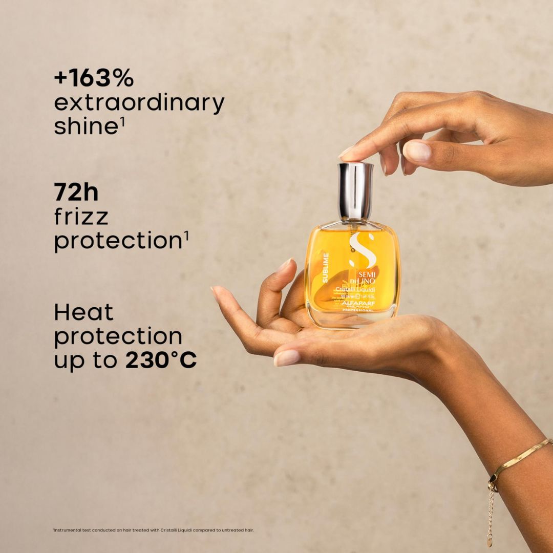 A hand holds a square glass bottle of hair oil with a silver cap, while another hand touches the top. The bottle contains a golden-yellow liquid and has white text including an 'S' logo.