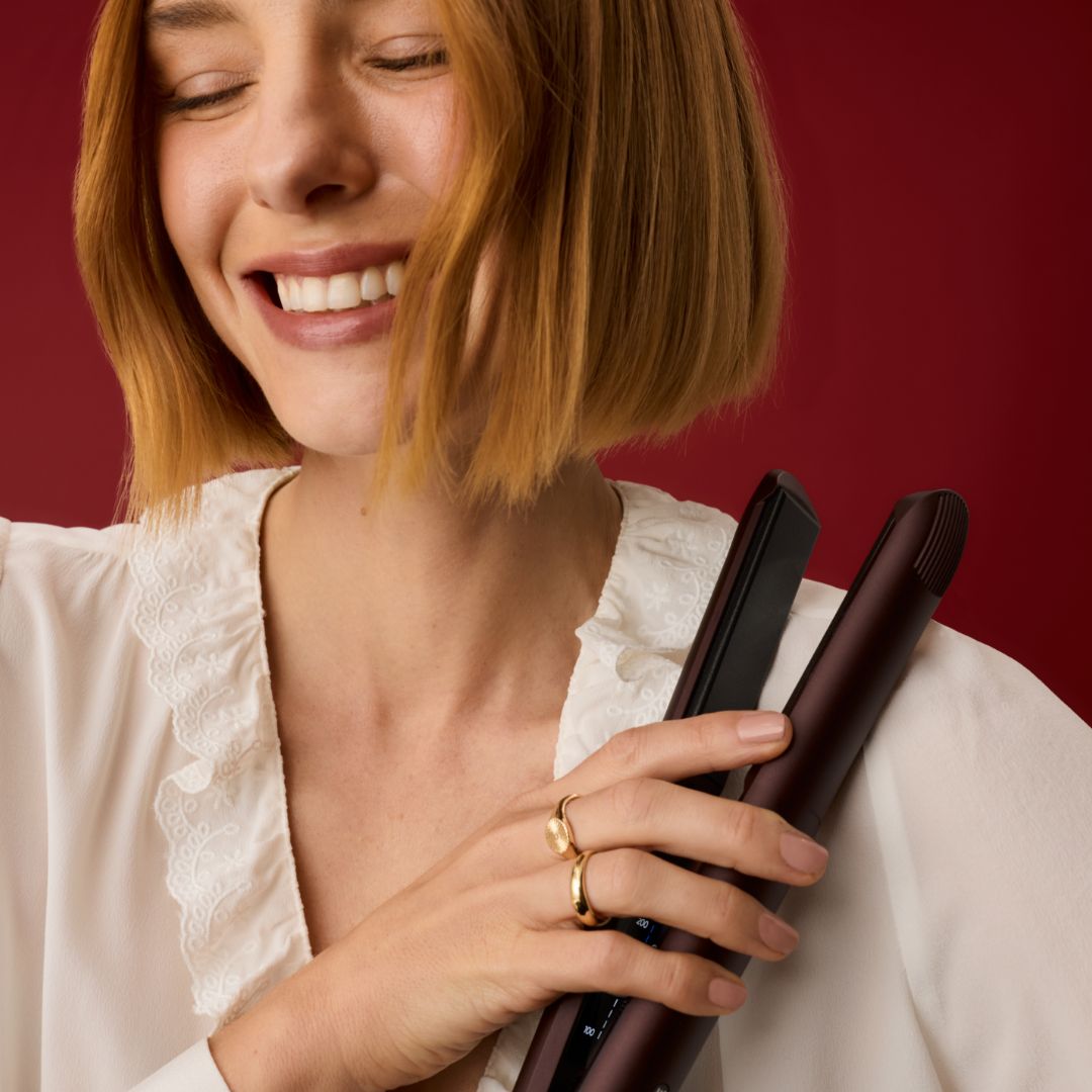 A woman with a short blonde bob holds a dark brown flat iron against her chest, wearing a white blouse and gold rings, set against a solid dark red background.