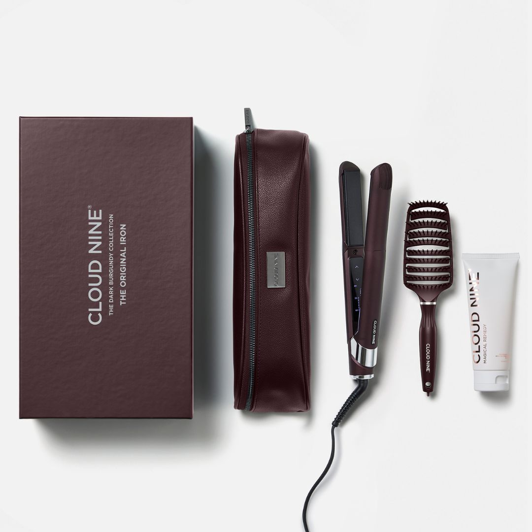 Cloud Nine hair styling set on a light gray background, including a dark brown presentation box, a matching zippered pouch, a flat iron, a barrel brush, and a small tube.