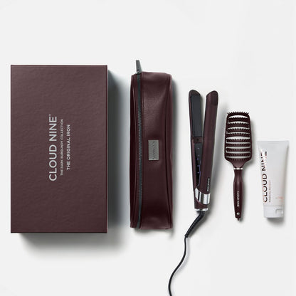 Cloud Nine hair styling set on a light gray background, including a dark brown presentation box, a matching zippered pouch, a flat iron, a barrel brush, and a small tube.