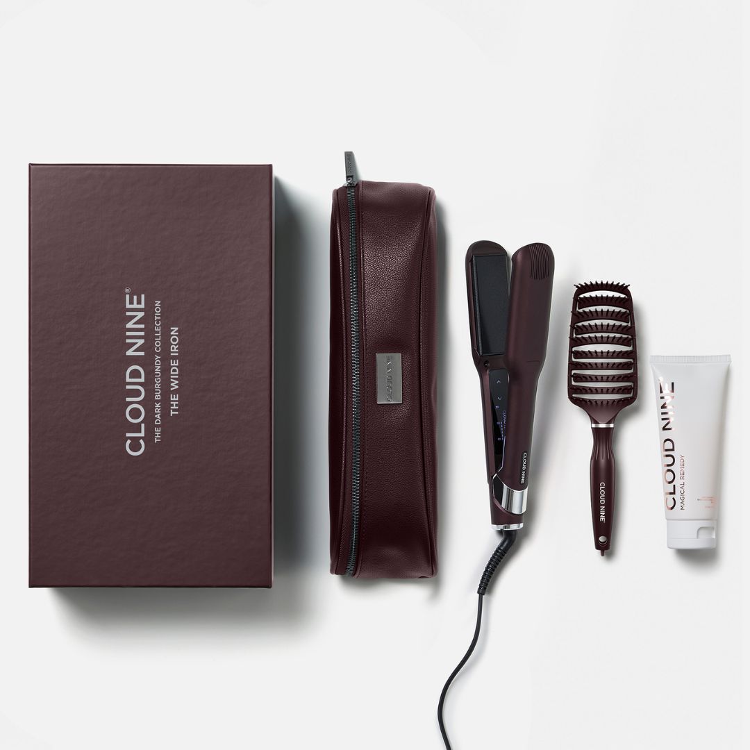 Cloud Nine The Wide Icon hair styling set on a light gray background, including a dark brown presentation box, a zippered case, a flat iron, a round brush, and a small tube.