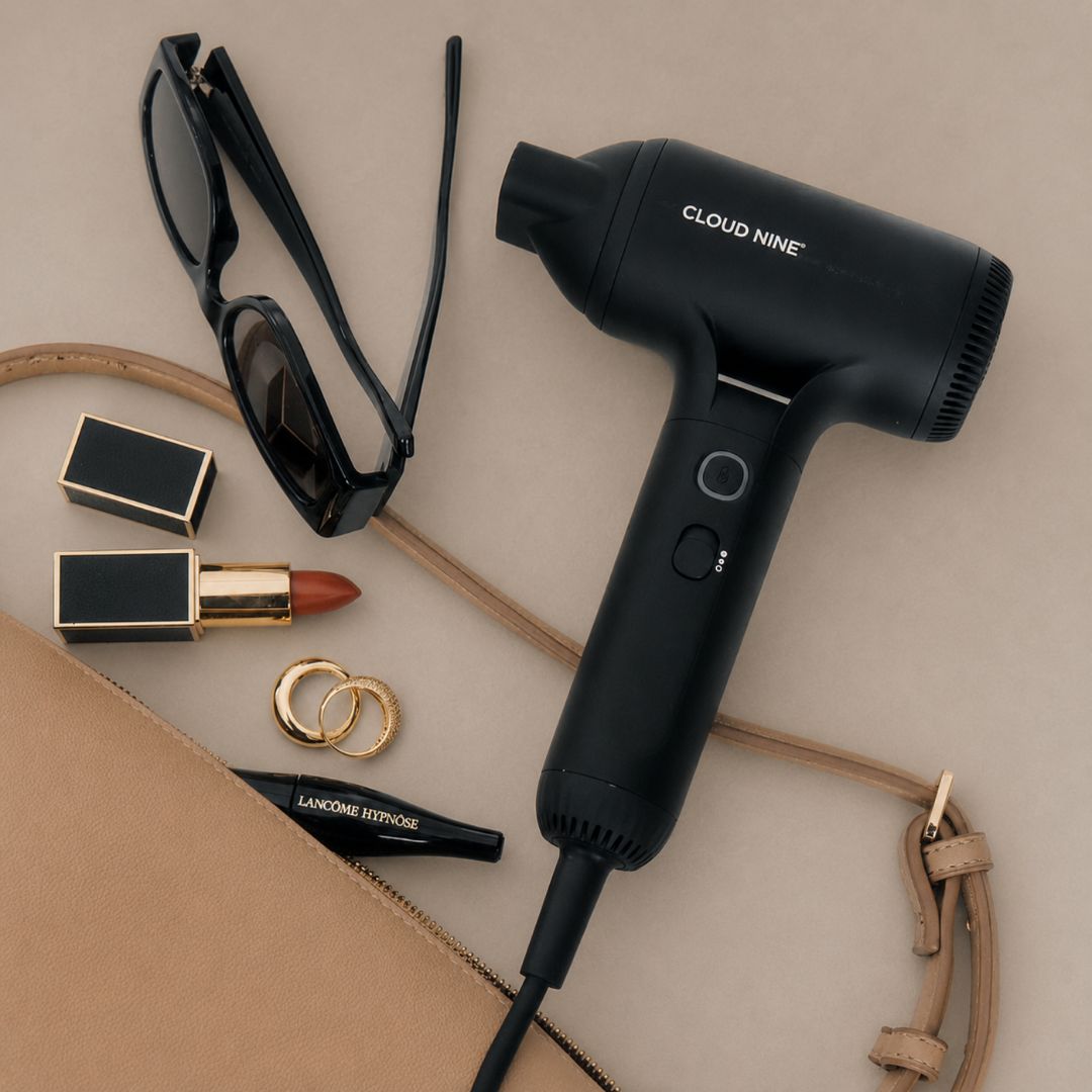 A black handheld hair dryer with the brand name 'CLOUD NINE' printed on the side, styled in a flat-lay arrangement with sunglasses, lipstick, rings, a black pencil, and a beige leather bag on a neutral beige background.