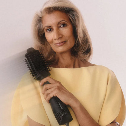A woman with shoulder-length blonde hair holds a black hair styling brush against a plain light background. She is wearing a pale yellow top.