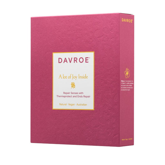 A rectangular pink product box for Davroe 'A lot of Joy Inside' shown at a slight angle on a plain white background. The front has a cream label with gold and dark pink text, and the side panel shows additional text and a small gold icon.