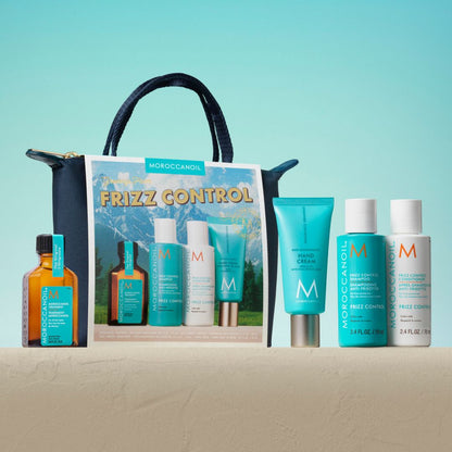 Moroccanoil Frizz Control gift set arranged in front of a branded tote bag on a beige surface against a turquoise gradient background. The set includes an amber bottle, a large turquoise hand cream tube, a turquoise leave-in conditioner bottle, a white treatment bottle, and a white shampoo bottle, with the tote bag behind them.
