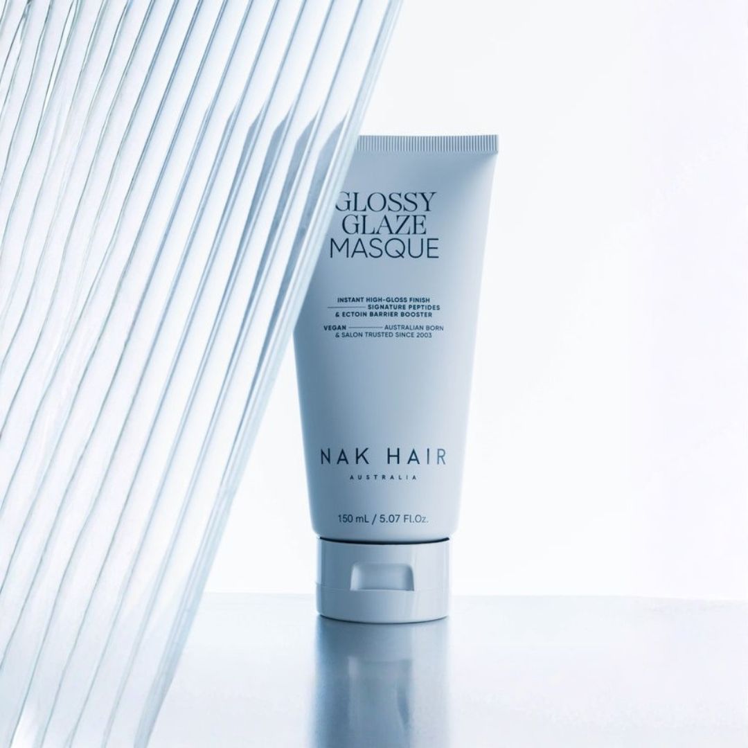 A tube of Nak Hair Glossy Glaze Masque hair conditioner standing upright on a reflective surface, with a modern ribbed glass panel on the left side of the frame.