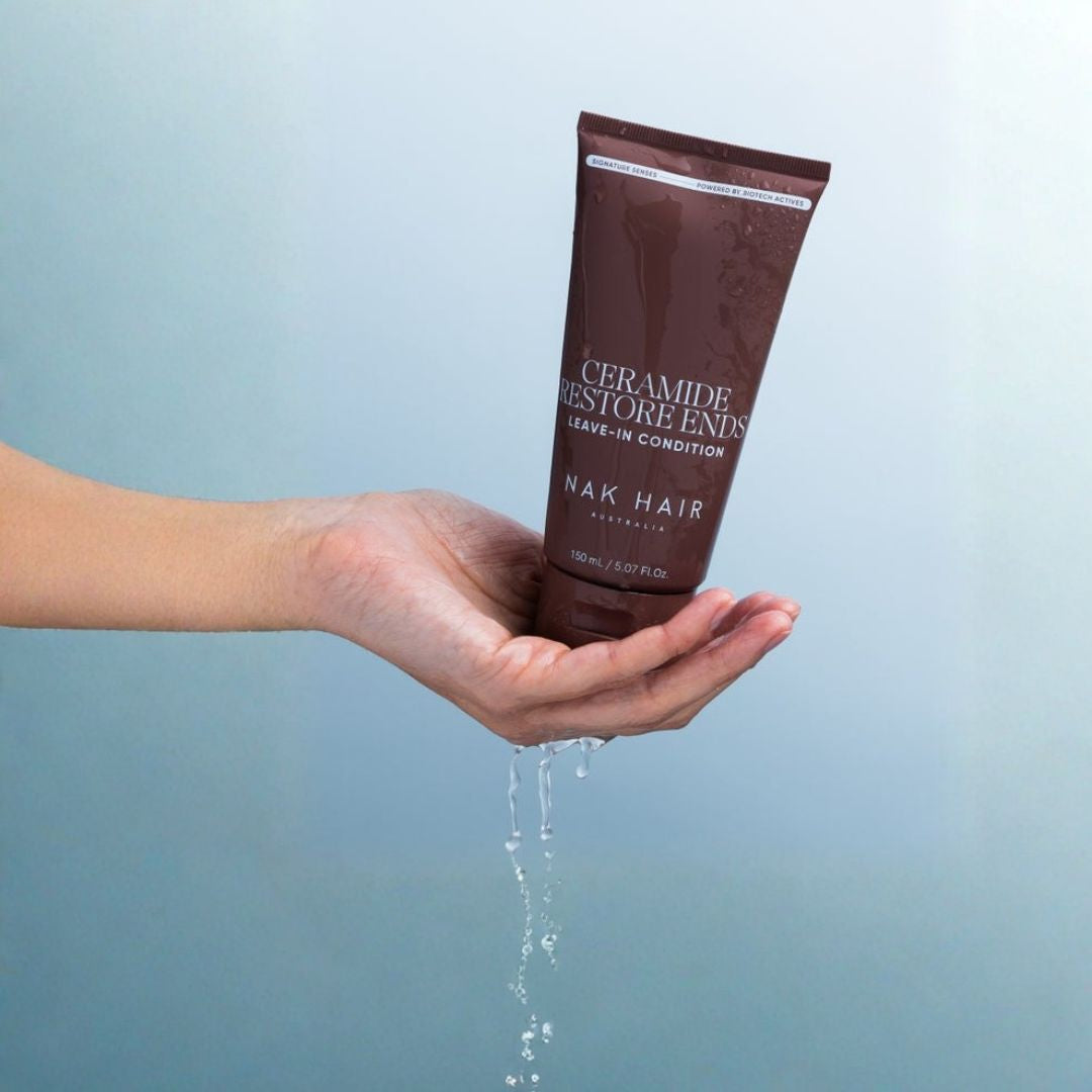 A hand holding a brown tube of NAK HAIR Ceramide Restore Evens Leave-In Conditioner, with water dripping from the bottom against a soft blue background.