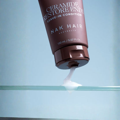 A dark brown squeeze tube of Nak Hair Ceramide Restore Ends Leave-In Condition is tilted over a clear glass surface, dispensing a white cream against a soft light blue background.