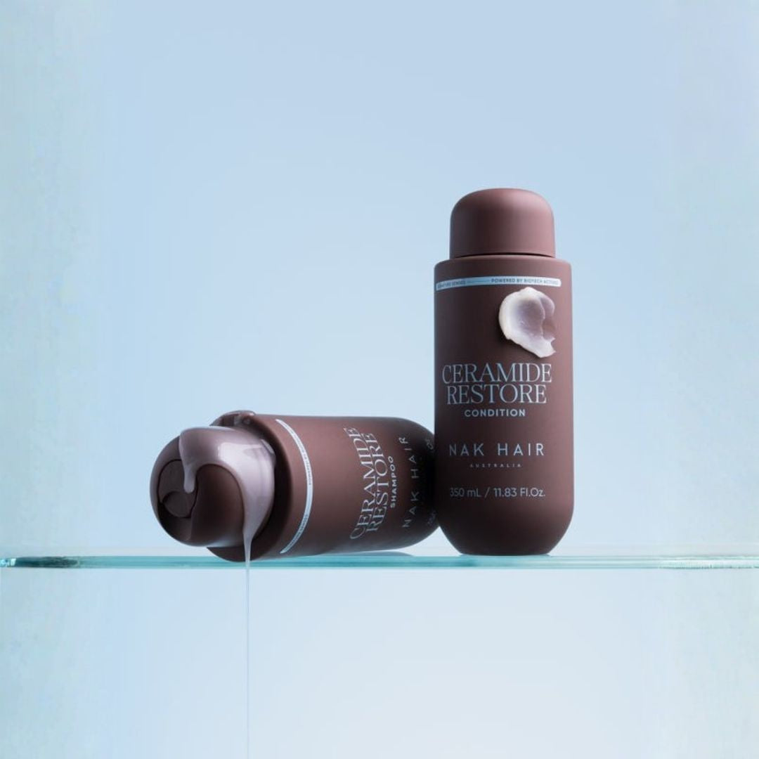Ceramide Restore Condition hair conditioner by NAK HAIR in a matte dark brown bottle, shown standing upright and lying on its side with a thin stream of the conditioner dripping from the open cap, set on a clear glass shelf against a light blue background.