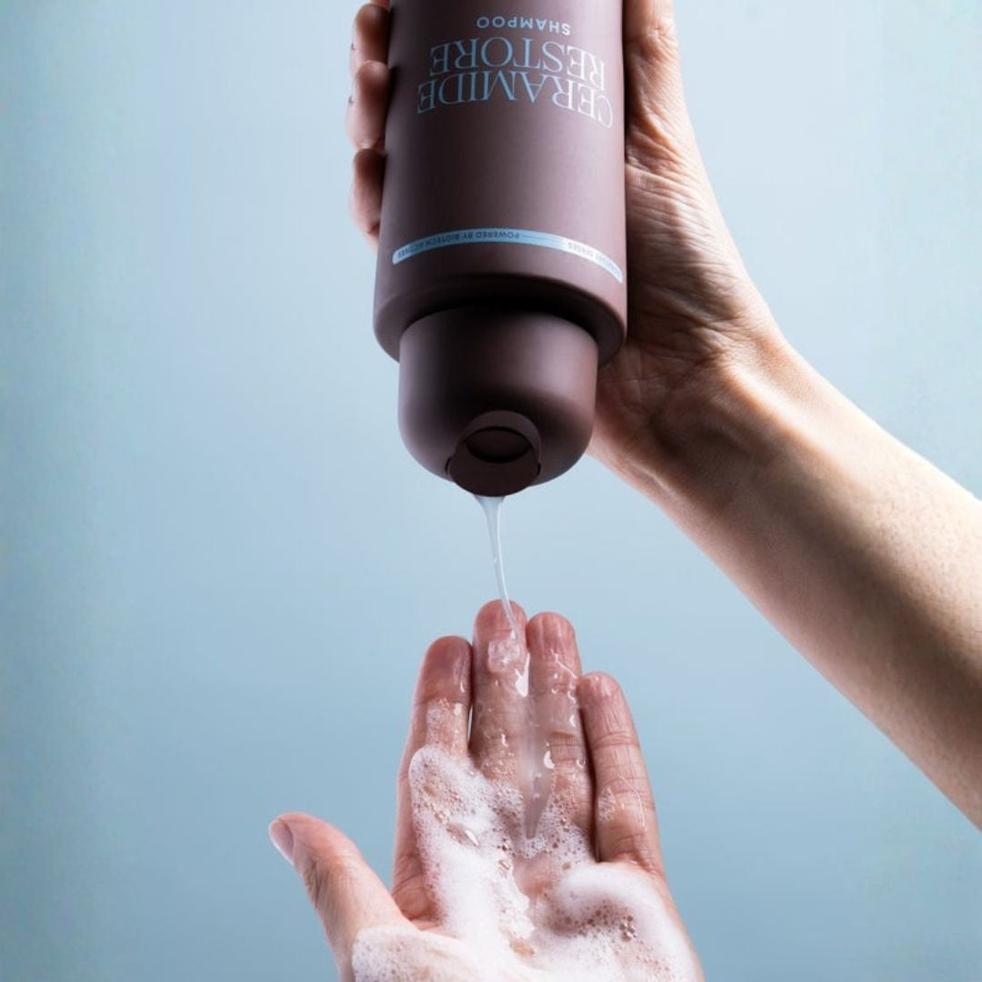 A hand holds a dark brown bottle of Ceramide Restore Shampoo, dispensing clear liquid onto the other hand which is covered in white foam, set against a solid light blue background.