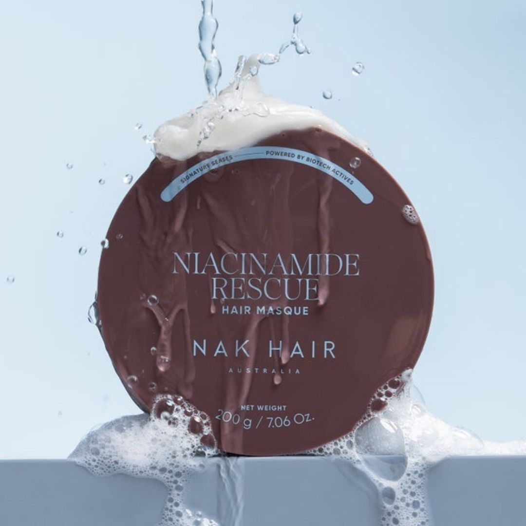 A round brown hair masque container with white text reading 'NIACINAMIDE RESCUE HAIR MASQUE' and 'NAK HAIR AUSTRALIA,' surrounded by splashing water and white foam against a pale blue background.