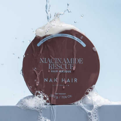 A round brown hair masque container with white text reading 'NIACINAMIDE RESCUE HAIR MASQUE' and 'NAK HAIR AUSTRALIA,' surrounded by splashing water and white foam against a pale blue background.