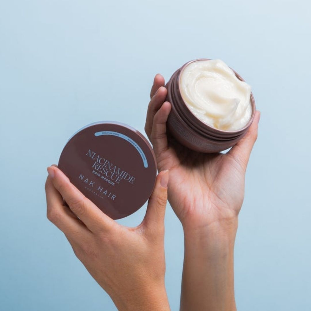 Two hands holding open jars of Nacnamide Rescued NAK Hair hair mask against a light blue background. One hand holds the dark brown lid showing the white text, and the other holds the open jar filled with a thick, off-white cream.