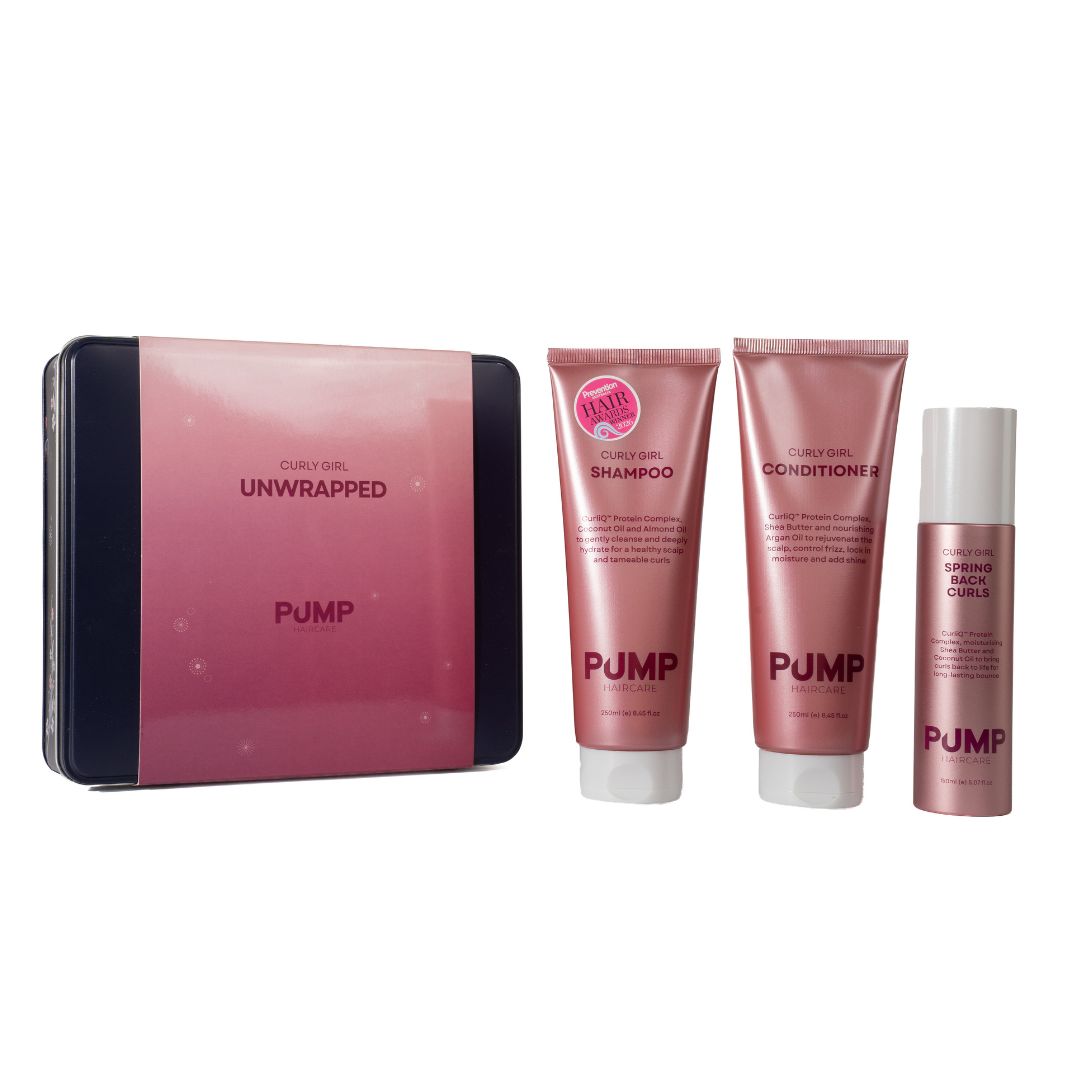 Pump Cosmetics Curl Hair Unwrapped set featuring a pink and black tin box alongside three matching pink haircare tubes: shampoo, conditioner, and a curl spray, all on a plain white background.