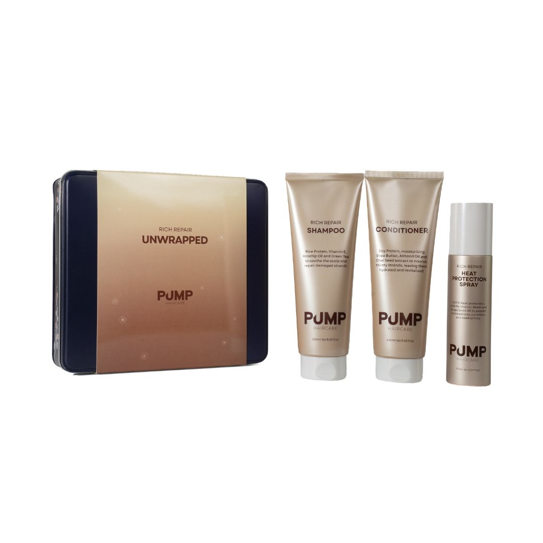 Pump Unwrapped bath and body set featuring a rectangular tin box and three matching squeeze tubes (shampoo, conditioner, and gel moisturizer) in a metallic champagne-gold color with dark navy blue side panels, on a plain white background.