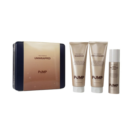 Pump Unwrapped bath and body set featuring a rectangular tin box and three matching squeeze tubes (shampoo, conditioner, and gel moisturizer) in a metallic champagne-gold color with dark navy blue side panels, on a plain white background.