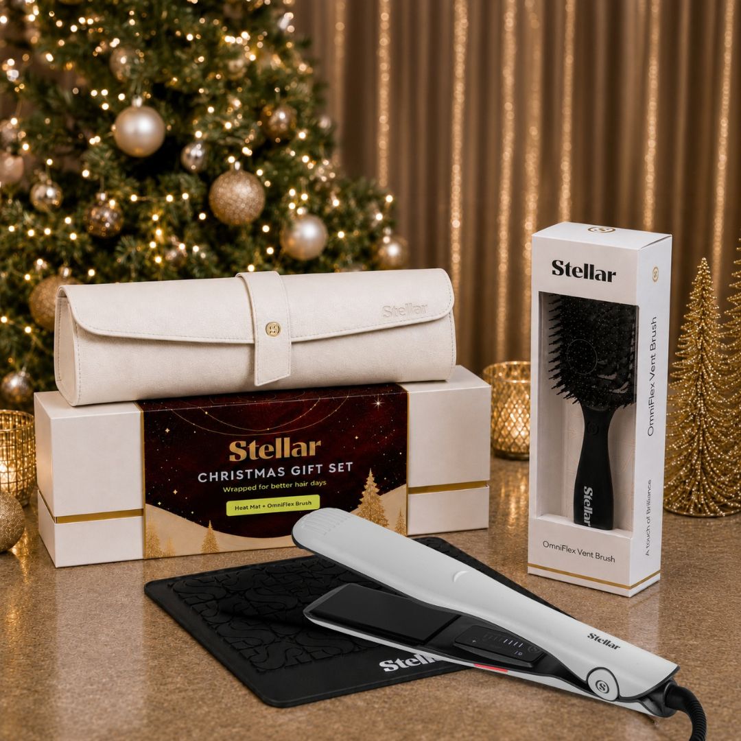 Stellar Christmas gift set featuring a white hair straightener, a black detangling brush in its box, and a cream-colored travel case on a tabletop in front of a decorated Christmas tree and gold curtains.