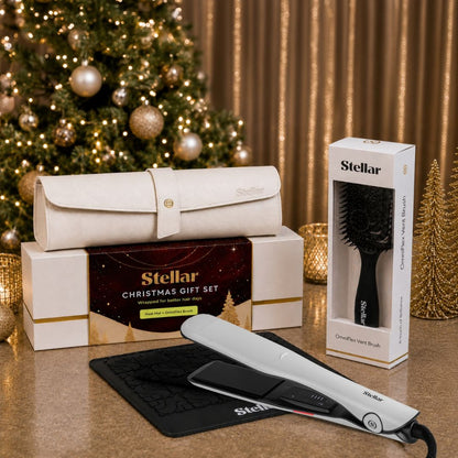 Stellar Christmas gift set featuring a white hair straightener, a black detangling brush in its box, and a cream-colored travel case on a tabletop in front of a decorated Christmas tree and gold curtains.