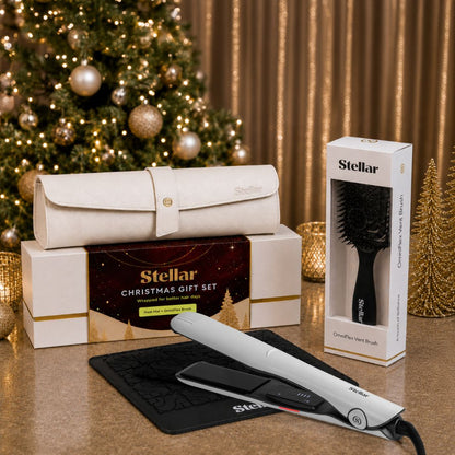 Stellar Christmas gift set featuring a white hair straightener, a black detangling brush in its box, and a cream-colored travel pouch resting on branded gift boxes, styled in front of a decorated Christmas tree and gold curtains.