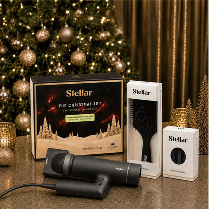 Stellar hair styling set displayed with its retail packaging on a tabletop in front of a decorated Christmas tree. The set includes a black hair dryer, a black hairbrush in a white box, and a black concentrator nozzle in a white box.
