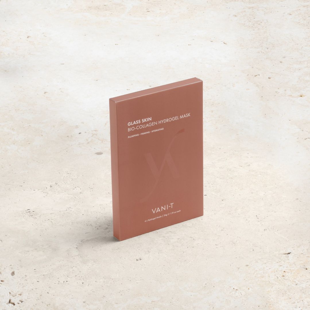 A rectangular box of VANI-T Glass Skin Bio-Collagen Hydrogel Mask in a terracotta pink color, standing upright on a light beige textured stone surface.