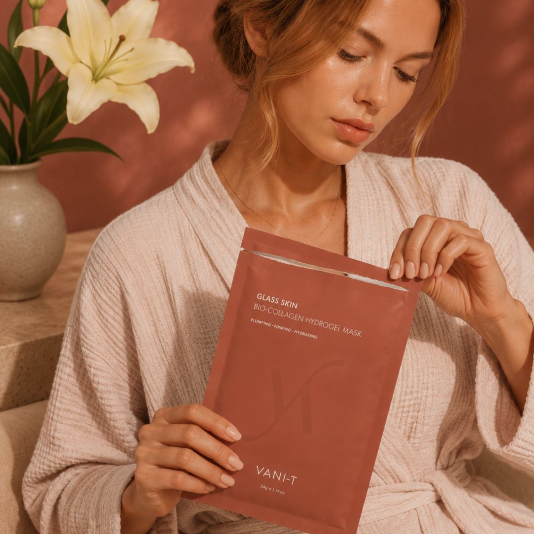 A woman in a beige waffle-knit robe holds a dusty rose pouch of Vani-T Glass Skin Bio-Collagen Hydrogel Mask. The background features a warm terracotta wall with leaf shadows and a vase of white lilies on a wooden table.