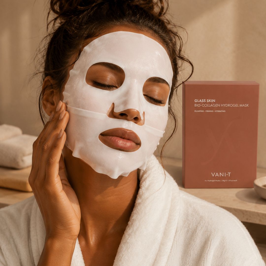 A woman wearing a white sheet mask and a white robe stands in a warm-toned bathroom setting next to a dusty rose box labeled 'GLASS SKIN BIO-COLLAGEN HYDROGEL MASK' by VANI-T.