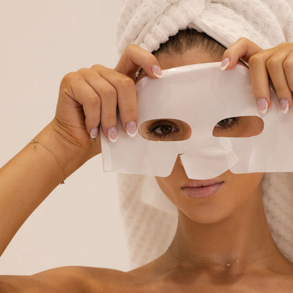 A woman with a white towel wrapped around her hair holds a white sheet mask in front of her face. She is wearing delicate gold jewelry, including a thin chain necklace and bracelet, against a clean, light beige background.