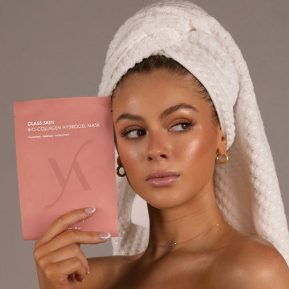 A woman with a white textured towel wrapped around her head holds a pink sheet mask package labeled 'GLASS SKIN BIO-COLLAGEN HYDROGEL MASK' against a plain taupe background.
