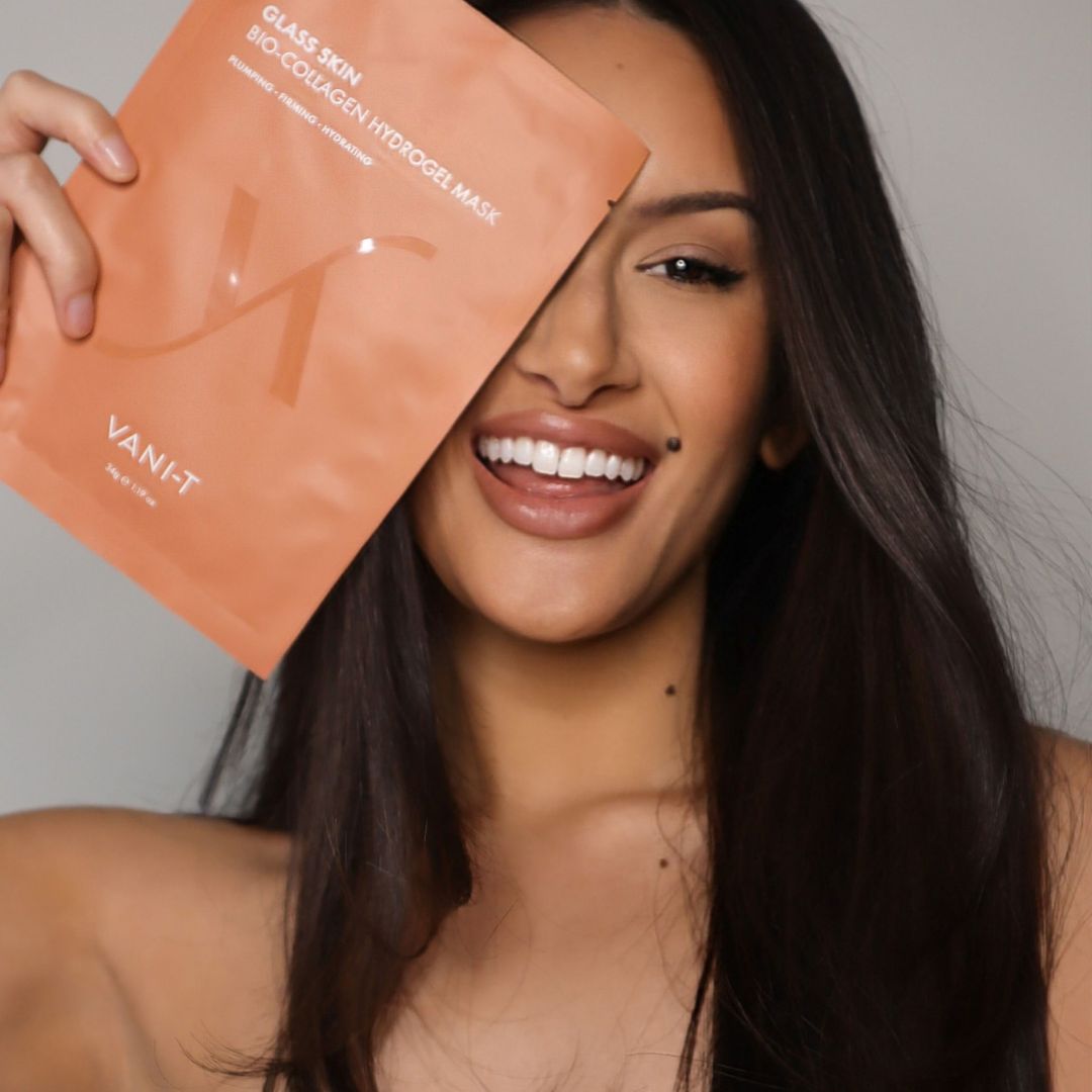 A woman with long dark hair holds a peach-colored Vani-T glass skin bio-collagen hydrogel mask pouch in front of her face against a neutral gray background.
