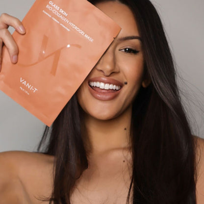 A woman with long dark hair holds a peach-colored Vani-T glass skin bio-collagen hydrogel mask pouch in front of her face against a neutral gray background.