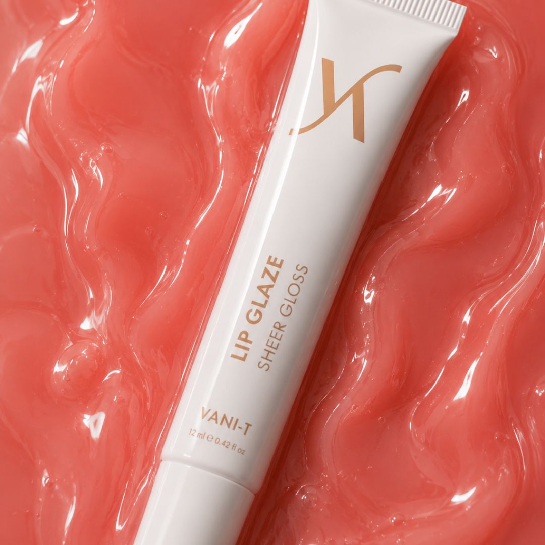 A white tube of Vani-T Lip Glaze Sheer Gloss with gold lettering, placed diagonally on a glossy, textured coral-pink background.