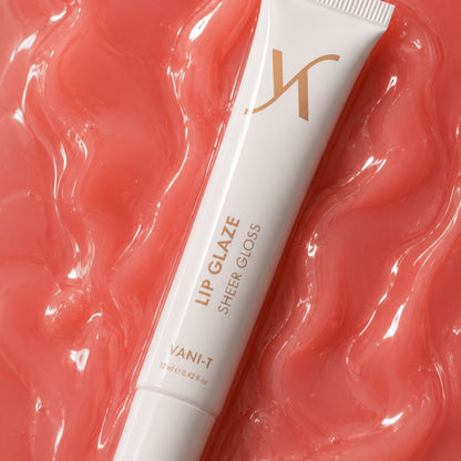 A white tube of Vani-T Lip Glaze Sheer Gloss with gold lettering, placed diagonally on a glossy, textured coral-pink background.