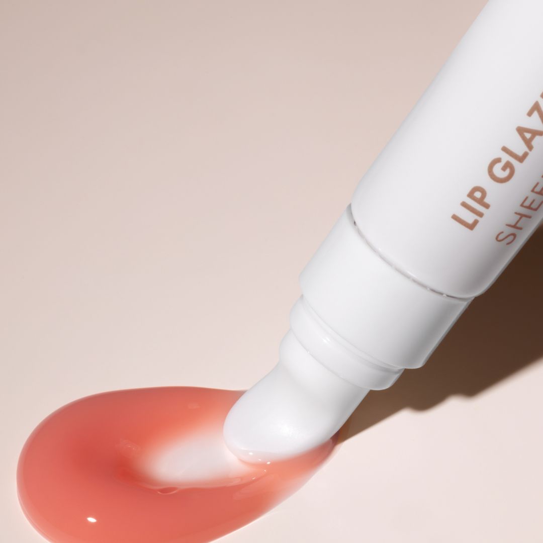 A close-up of a white lip gloss tube dispensing a glossy peach-pink lip gloss onto a soft beige background. The tube shows partial text reading 'LIP GLAZ.