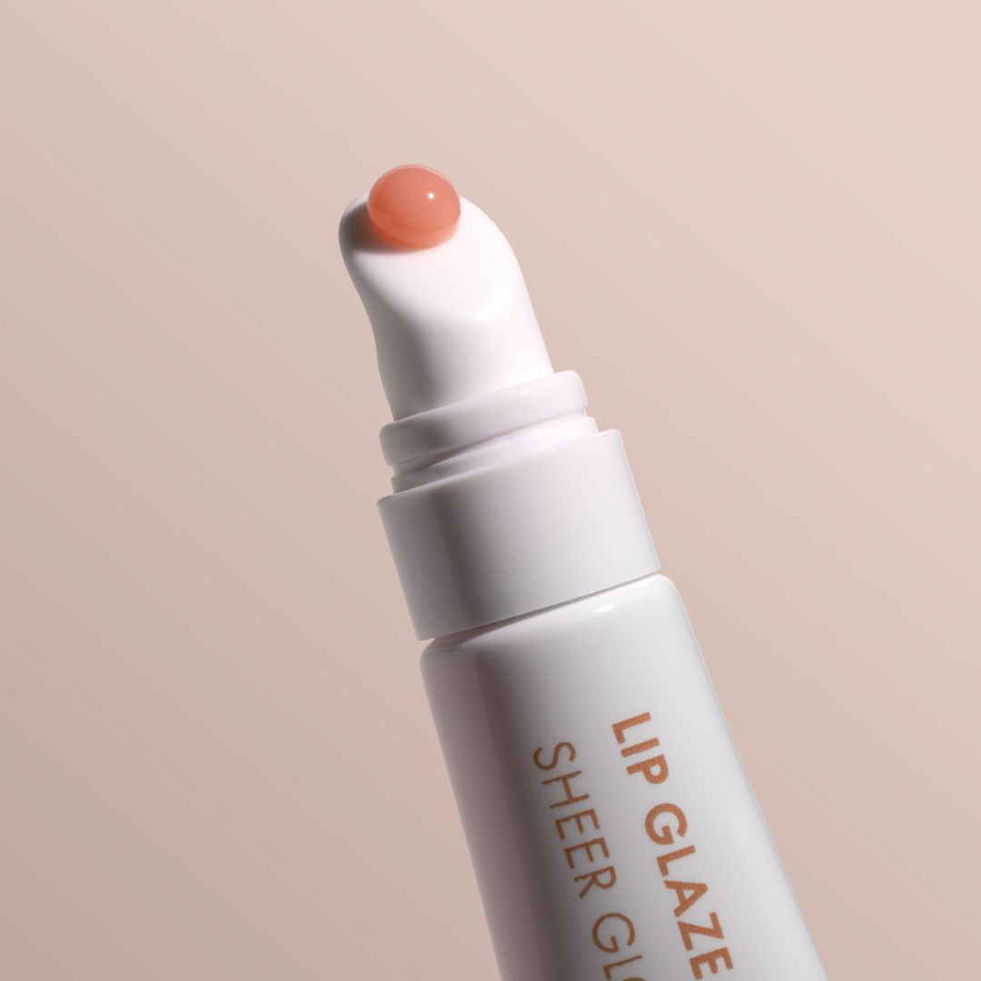 A close-up of a white lip glaze tube with a rounded applicator tip holding a peach-colored gloss, set against a solid pale beige background. The tube features orange text reading 'LIP GLAZE SHEER GLAZE'.