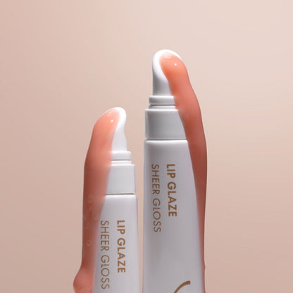 Two white lip gloss tubes with clear caps, one larger and one smaller, showing a sheer peach-pink gloss. The larger tube has a finger pressing the clear cap, and both tubes are labeled 'LIP GLAZE SHEER GLOSS' on a soft beige background.