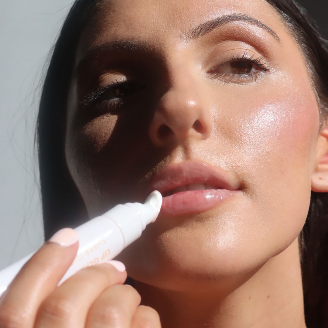 Close-up of a woman applying a white lip balm or gloss stick to her lips in bright sunlight.