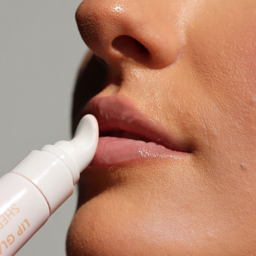 Close-up of a person applying a glossy lip balm or lip gloss from a white tube with a rounded applicator tip to their lips.