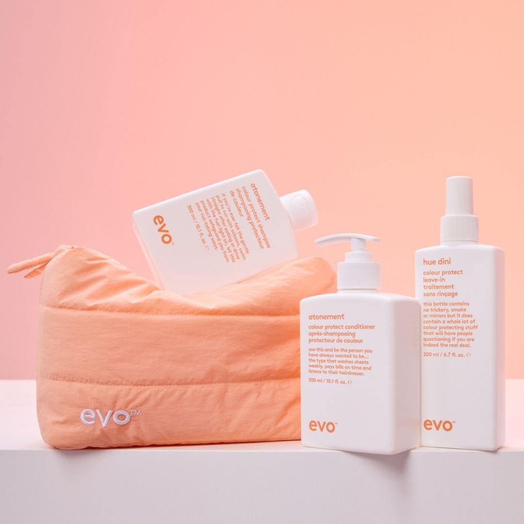 Three EVO haircare products arranged on a white surface against a soft peach-pink background: a peach zip pouch with the EVO logo, a white bottle tilted behind the pouch, and two white pump/spray bottles in front with orange text. The pouch and bottles are the products being shown.